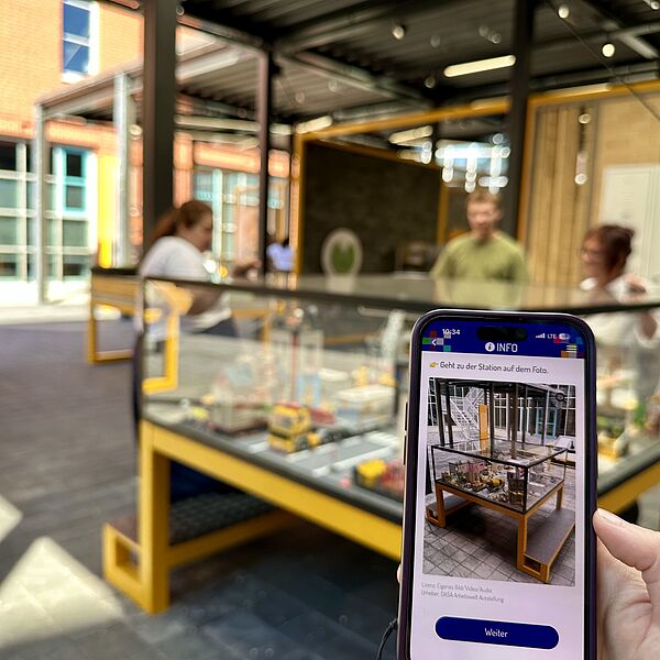 Smartphone mit Ansicht der App und im Hintergrund eine Vitrine in der Ausstellung Hochbau