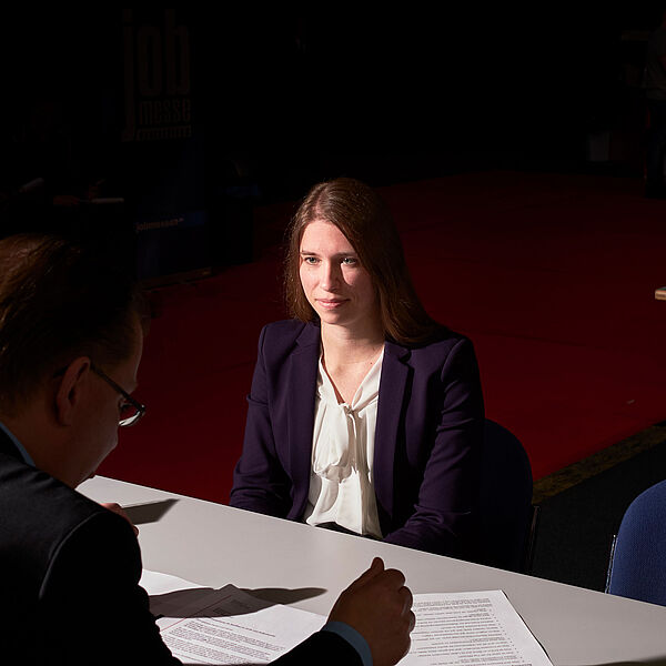 Eine junge Frau sitzt einem Mann an einem Schreibtisch bei einem Bewerbungsgespräch gegenüber. 