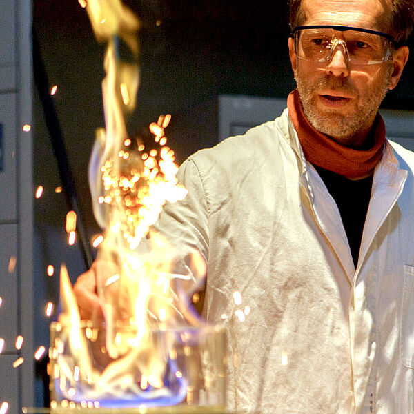 Ein Mann tr&auml;gt ein Laborkittel und f&uuml;hrt ein Experiment mit Feuer vor.