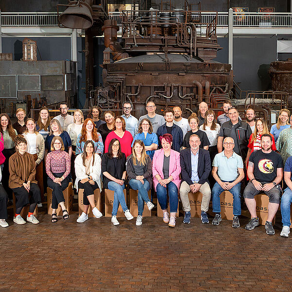 Das Team der DASA steht und sitzt in einem Halbkreis in der Stahlhalle der DASA