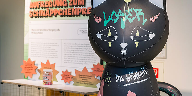 Katzen-Skulptur vor einer Texttafel in der Ausstellung "Alles im Angebot"