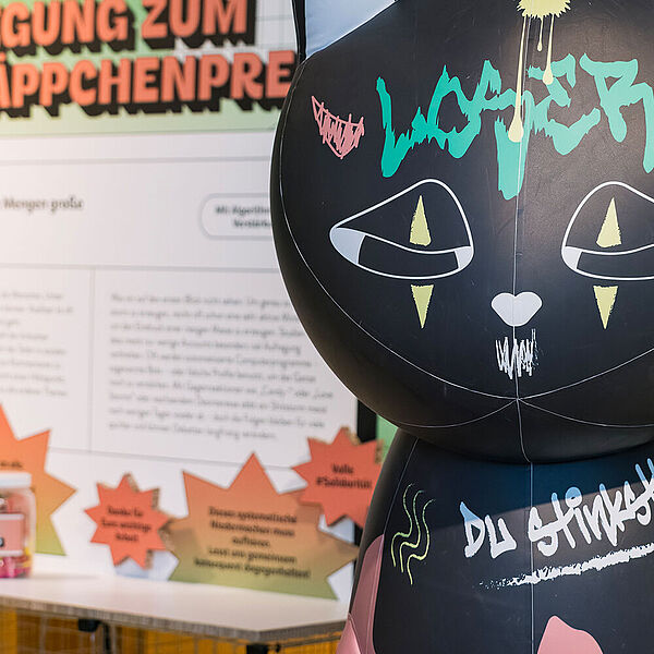 Katzen-Skulptur vor einer Texttafel in der Ausstellung "Alles im Angebot"