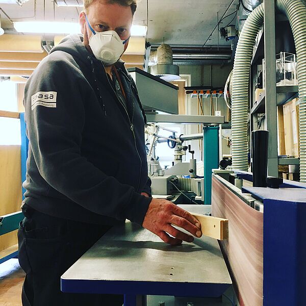 Ein Kollege aus der Tischlerei steht an einer Maschine und trägt dabei einen Mundschutz