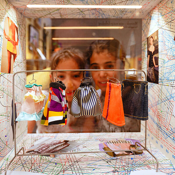 Zwei Mädchen schauen in eine Box in der Miniaturkleider auf einem Bügel hängen.