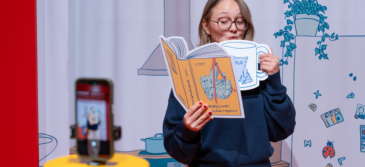 Einschlafen Frau hat eine große Teetasse und ein Buch in der Hand, davor ist ein Handy zu sehen. Das ist ein Selfie-SPot in der Ausstellung "GUte Nacht".